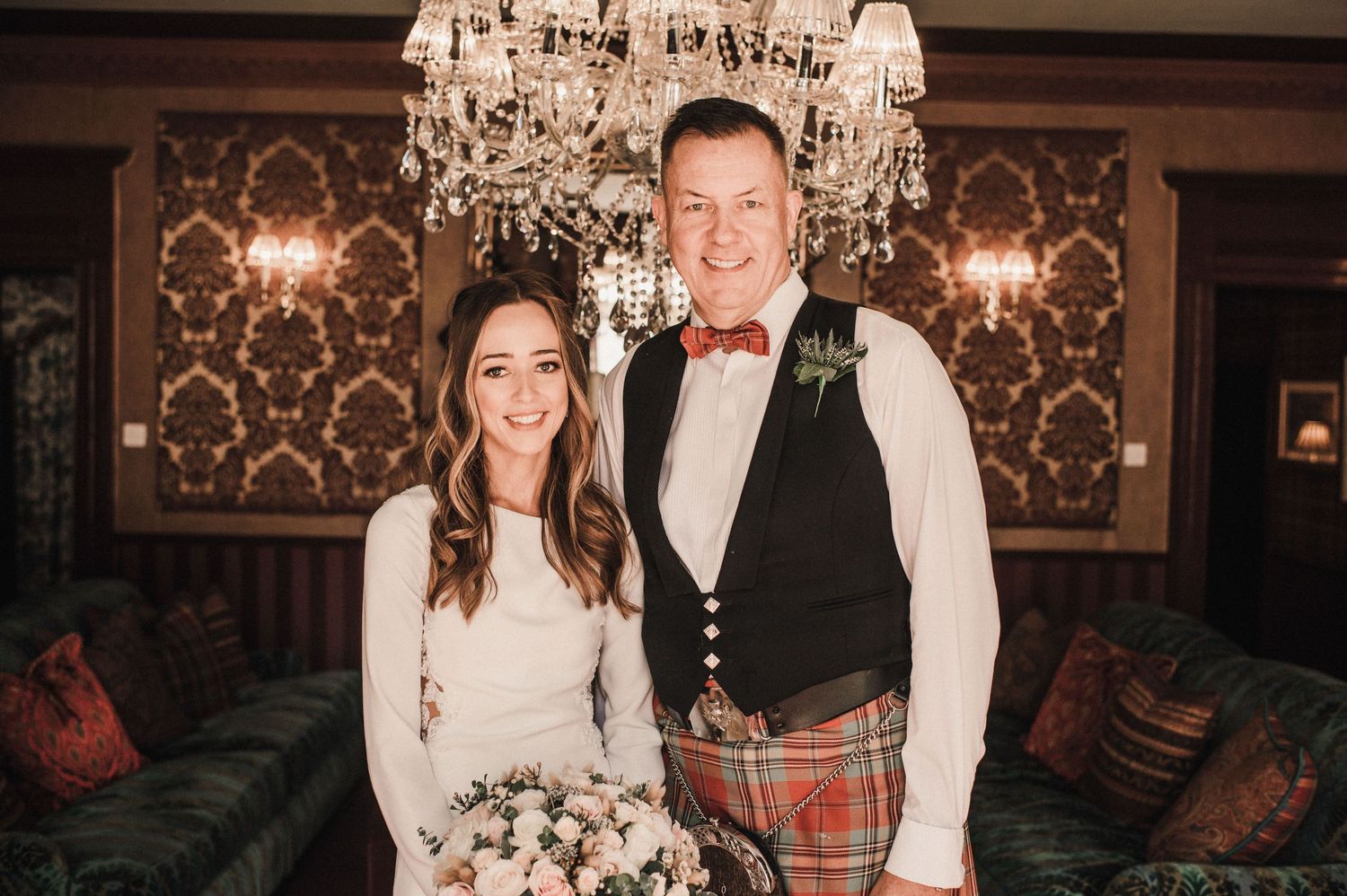 Newlyweds pose under chandelier with groom in traditional Scottish kilt and bride in white dress.