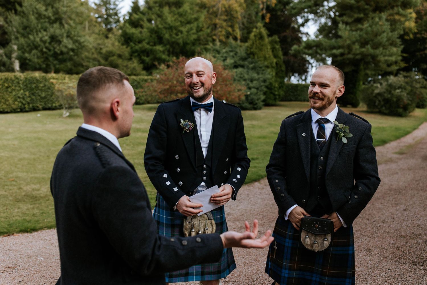 Groomsmen in traditional Scottish attire interact on garden grounds.