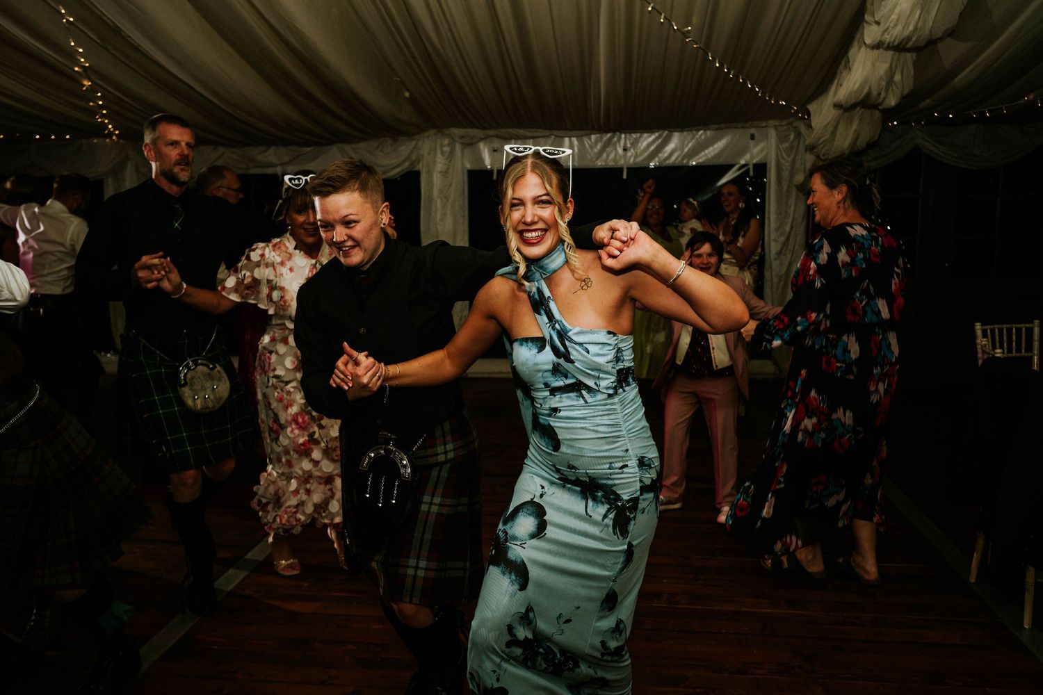 Wedding party dancing and having fun under a white tent with decorative lighting.