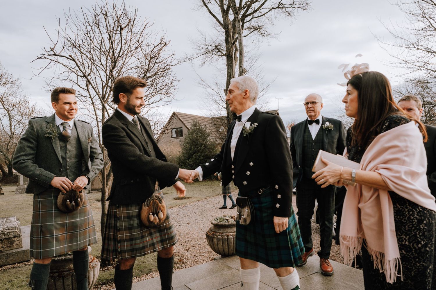 Wedding guests in traditional Scottish kilts gathering outside a stone church.