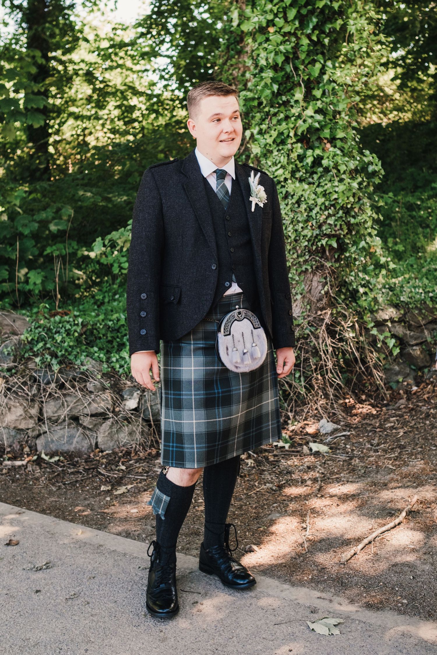 A person wearing traditional Scottish formal wear with kilt and sporran poses on a stone path.