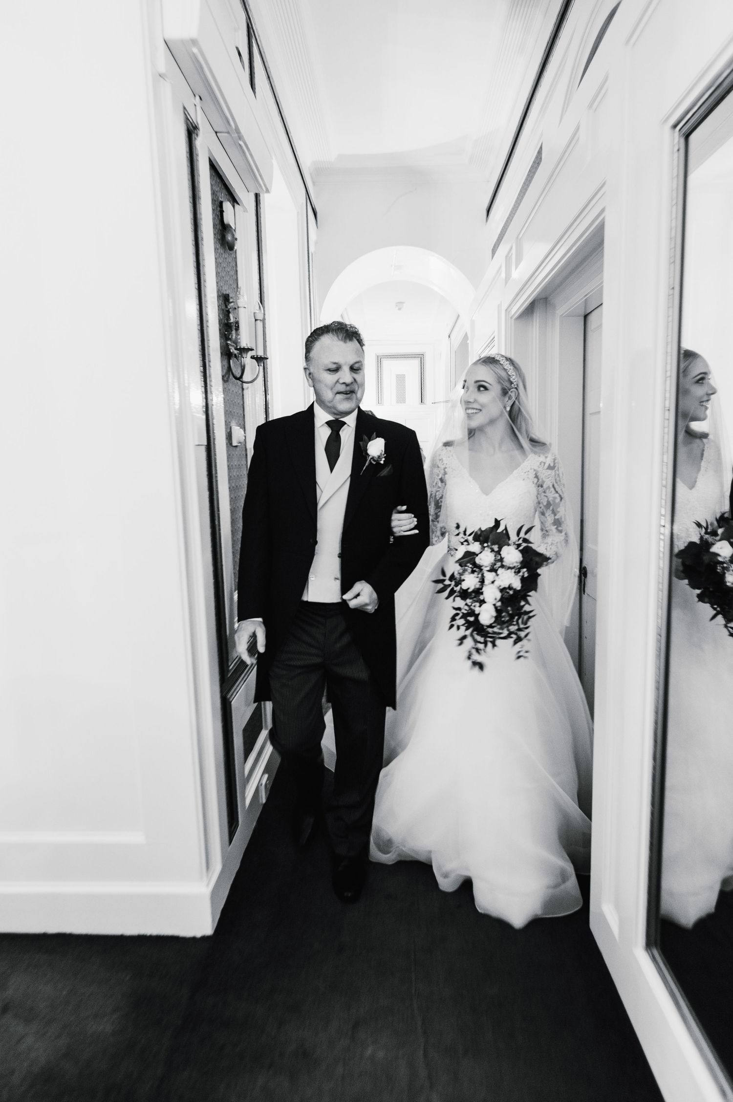 A black and white photo of a wedding party walking down a hallway lined with mirrors.