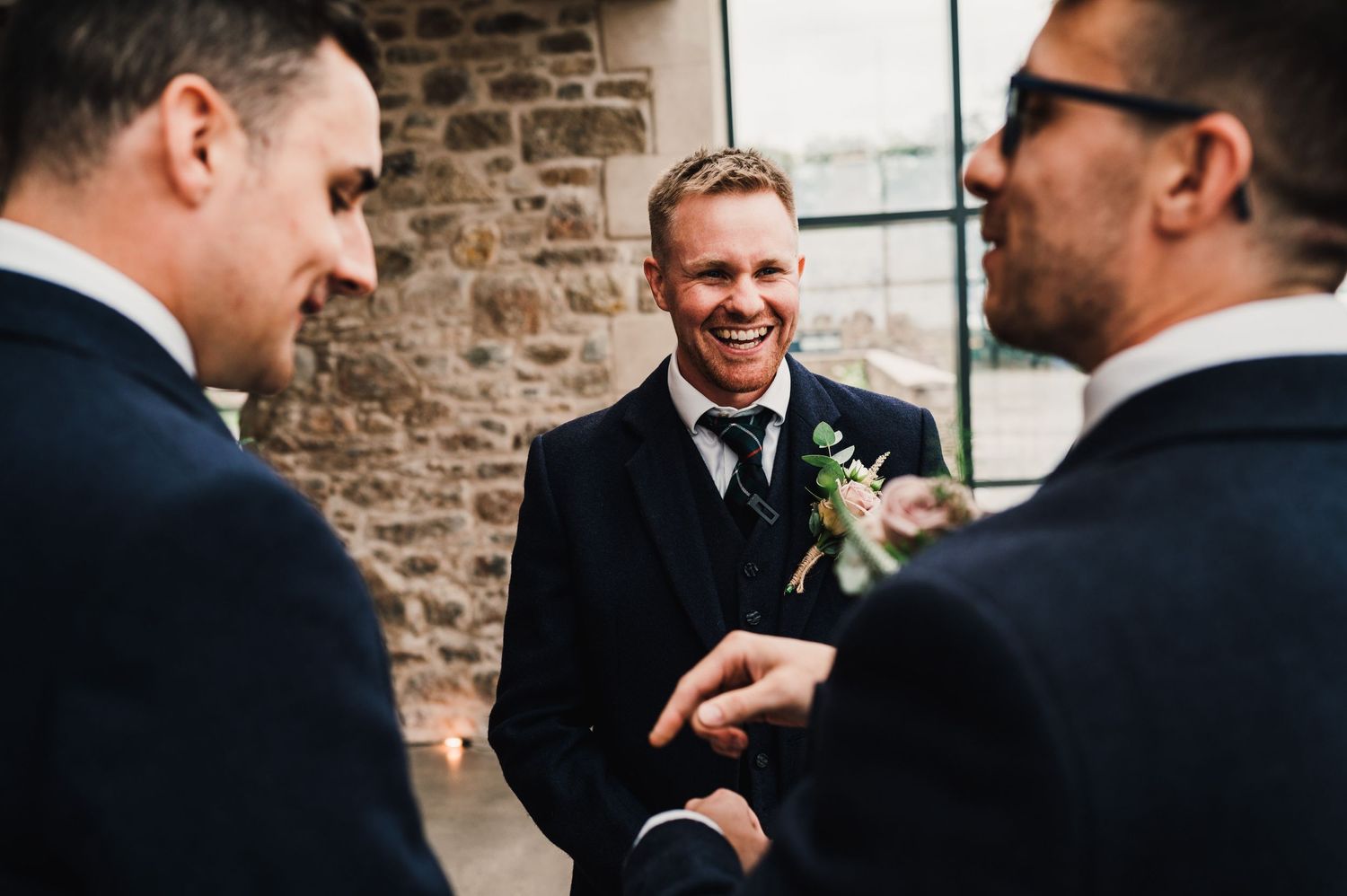 Groomsmen sharing a lighthearted moment before the wedding ceremony in a stone-walled venue.
