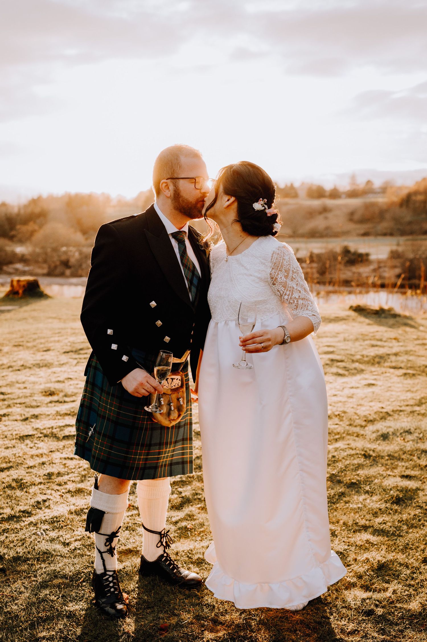 A Scottish wedding couple embraces in golden sunlight, with the groom wearing a traditional kilt and the bride in lace.