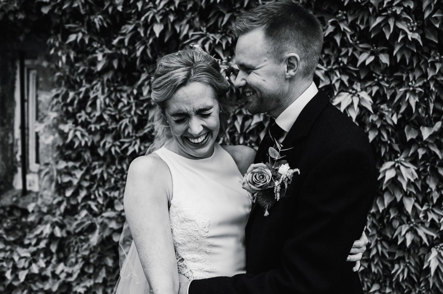 Black and white candid photo of newlyweds laughing together against a wall of greenery.