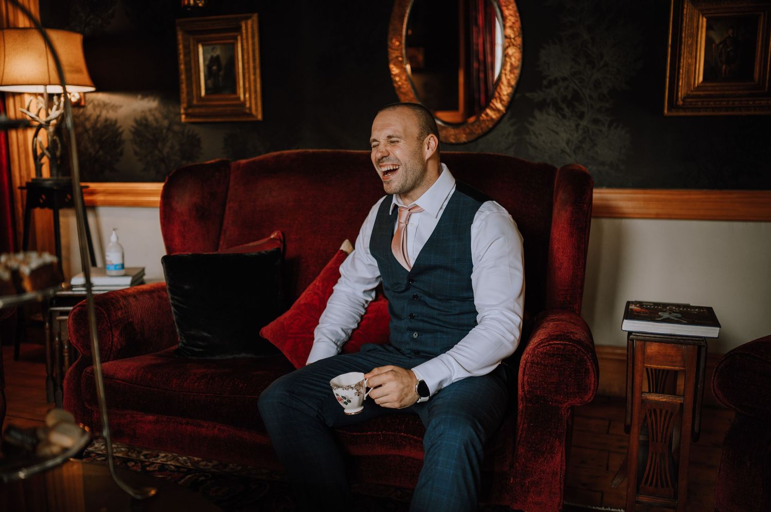 Person in vest and dress shirt laughing while seated on red vintage couch.