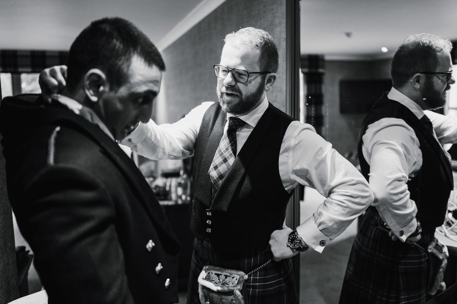 Men in formal attire having a conversation in black and white at an indoor gathering.