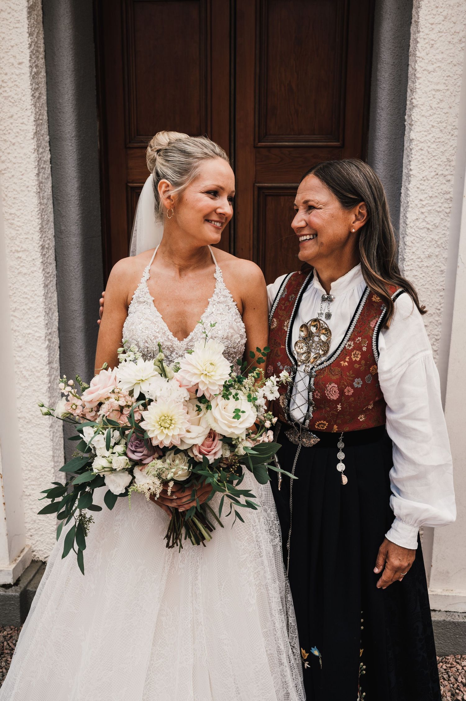 A bride in a white lace dress stands with someone wearing traditional Norwegian folk costume at a wedding.