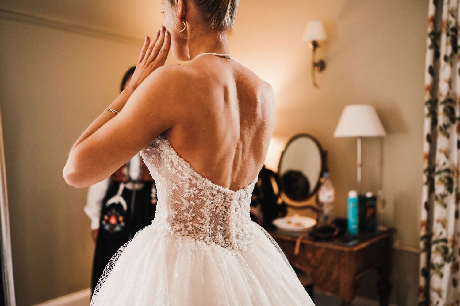 Back view of a wedding dress showing intricate lace corset detail and tulle skirt in a warmly lit dressing room.