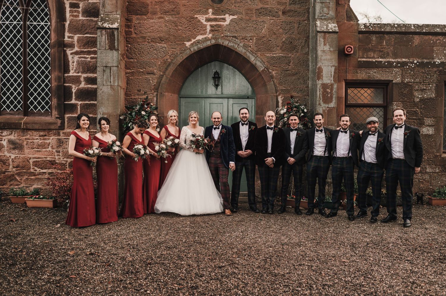 Wedding party with bridesmaids in red and groomsmen in black at stone church entrance.