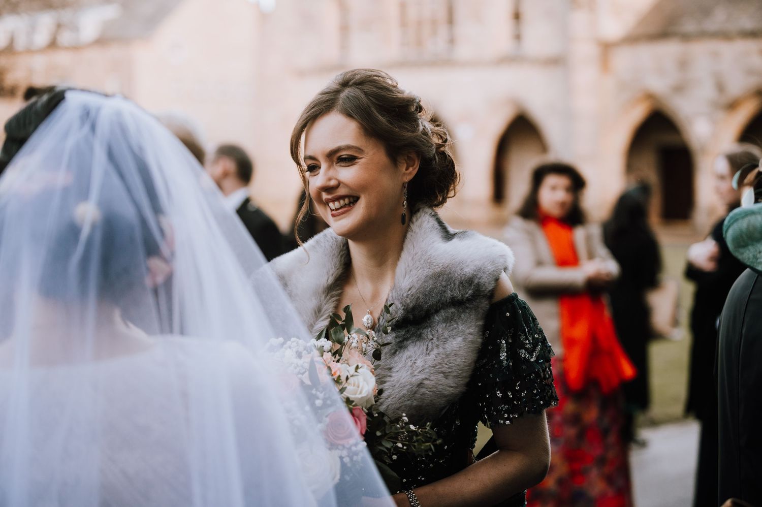 A guest wearing a fur stole smiles while holding flowers at a winter wedding celebration.