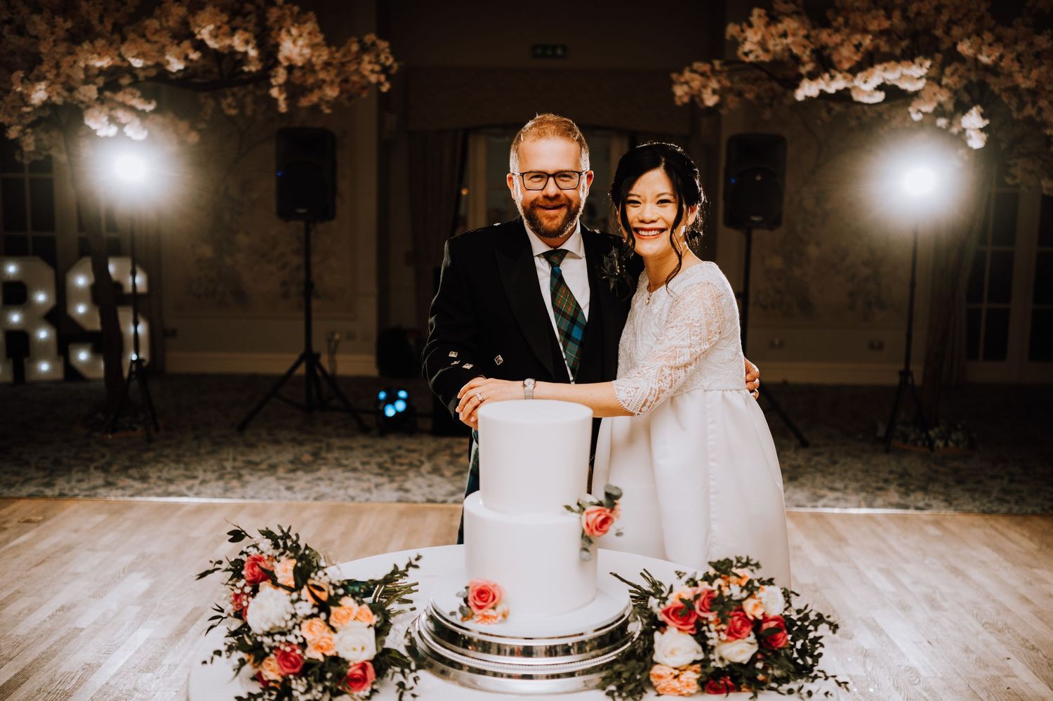 A couple cuts their three-tier white wedding cake surrounded by pink floral decorations.