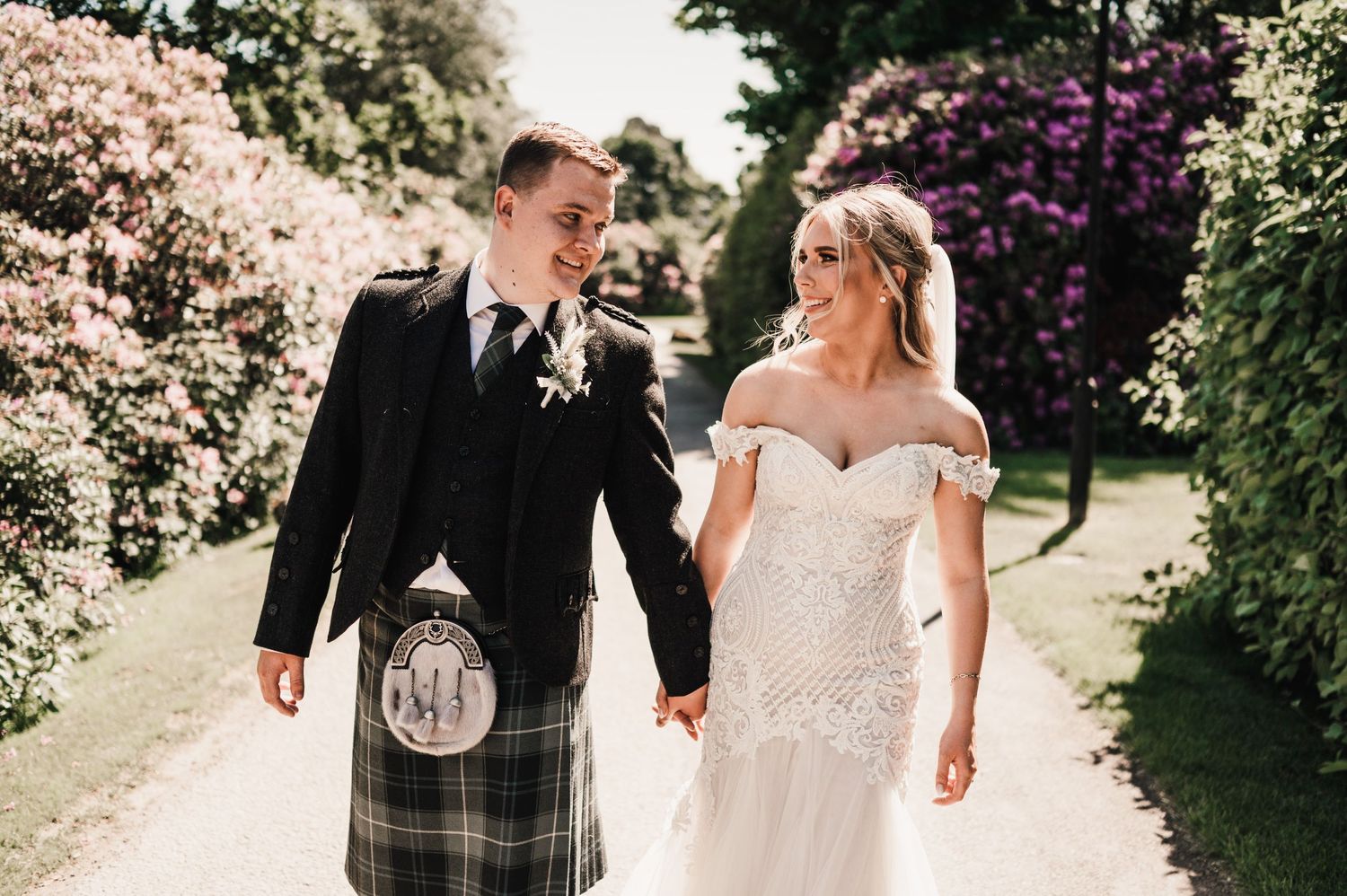 A couple strolls through a garden path lined with pink and purple flowering bushes.