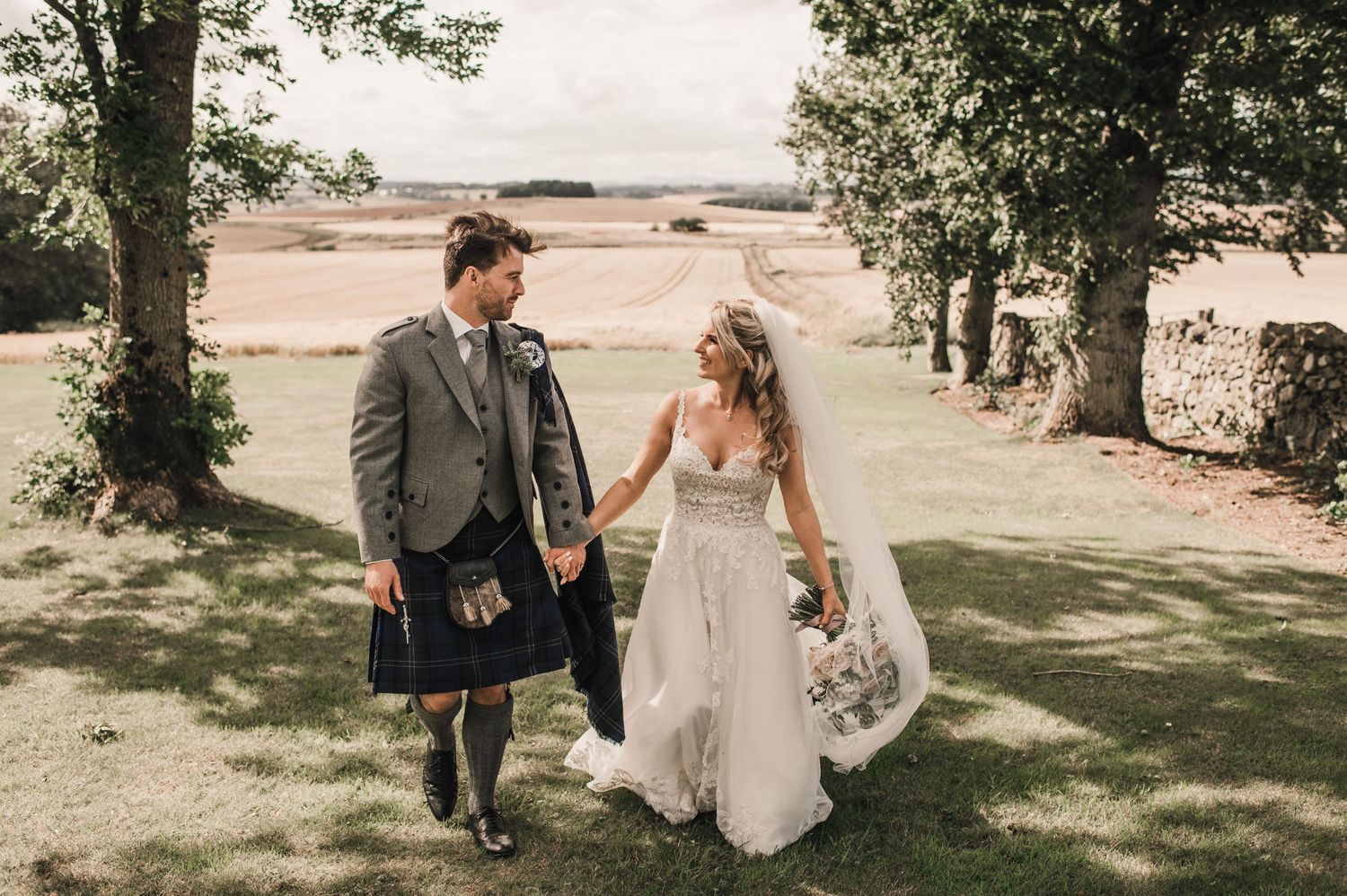 A bride and groom in traditional Scottish attire walk together through a sunlit garden.
