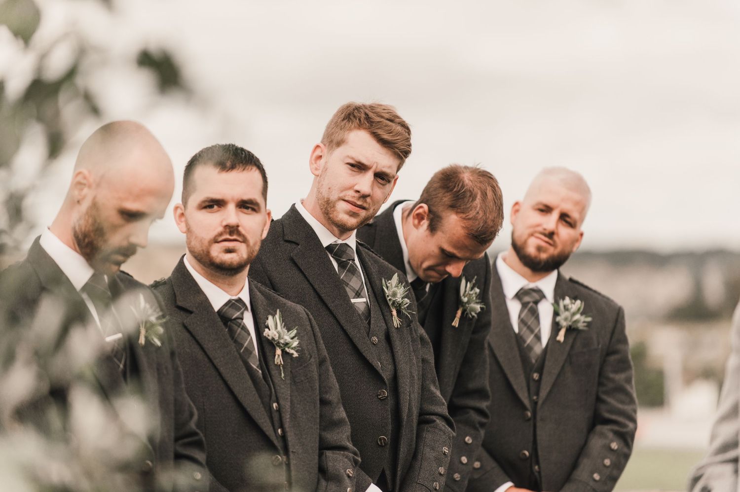 A group of groomsmen in dark suits standing together outdoors on a cloudy day.