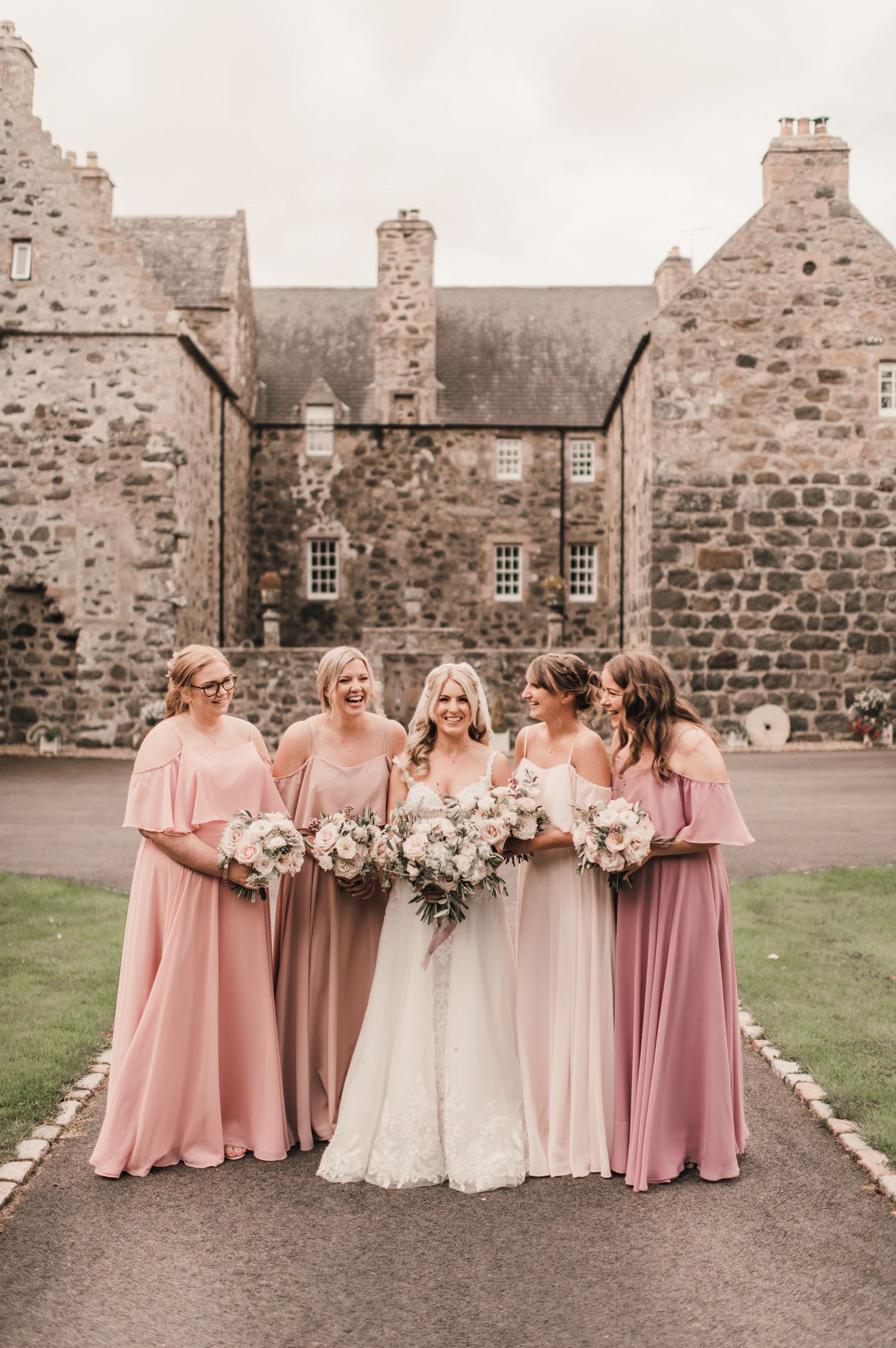 Bridal party in flowing pink dresses posing for photos outside a historic Scottish stone castle.