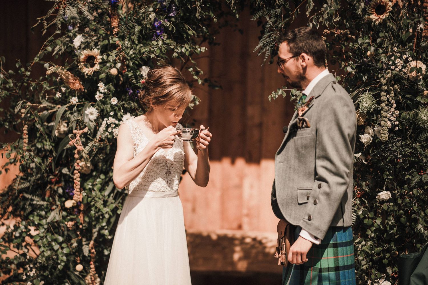 A romantic wedding moment with Scottish traditional dress under a floral arch in warm sunlight.
