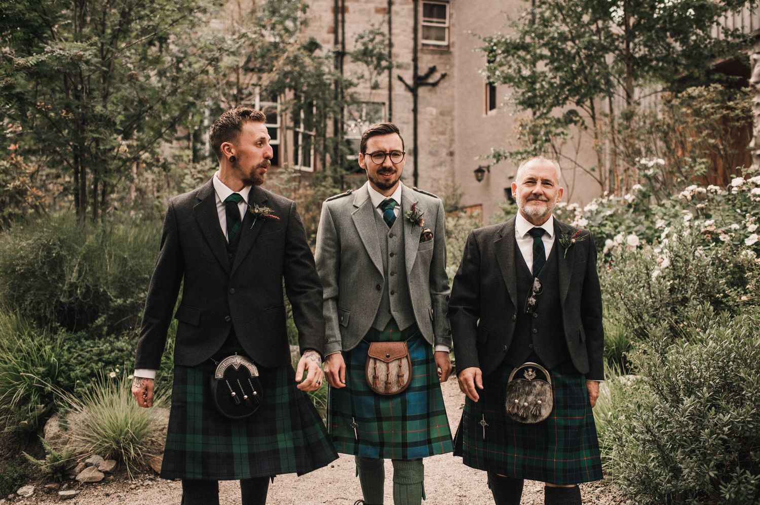 Three people wearing traditional Scottish kilts and formal jackets standing outdoors.
