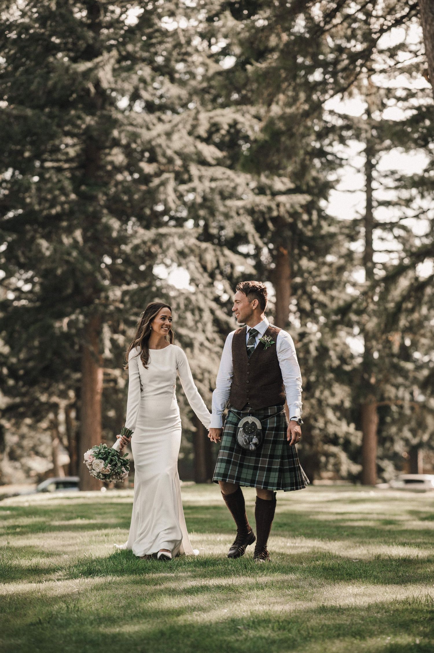 Wedding couple walks hand in hand on grass, with groom in traditional Scottish kilt and bride in elegant white dress.