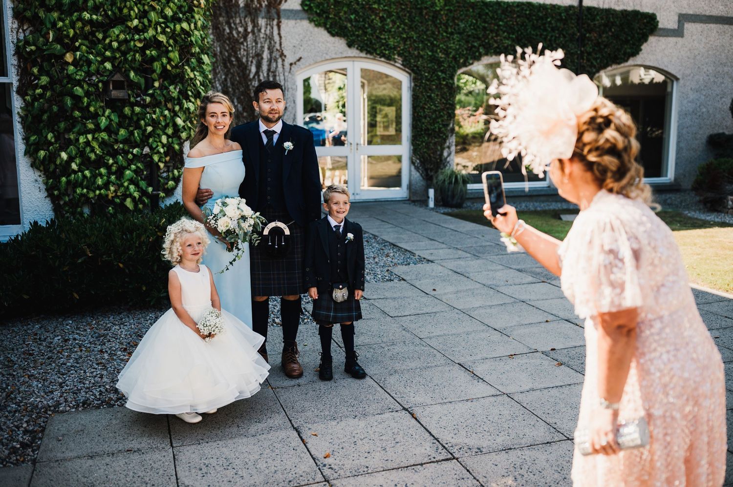 Wedding scene with flower girl in white dress and kilted guests being photographed outdoors.