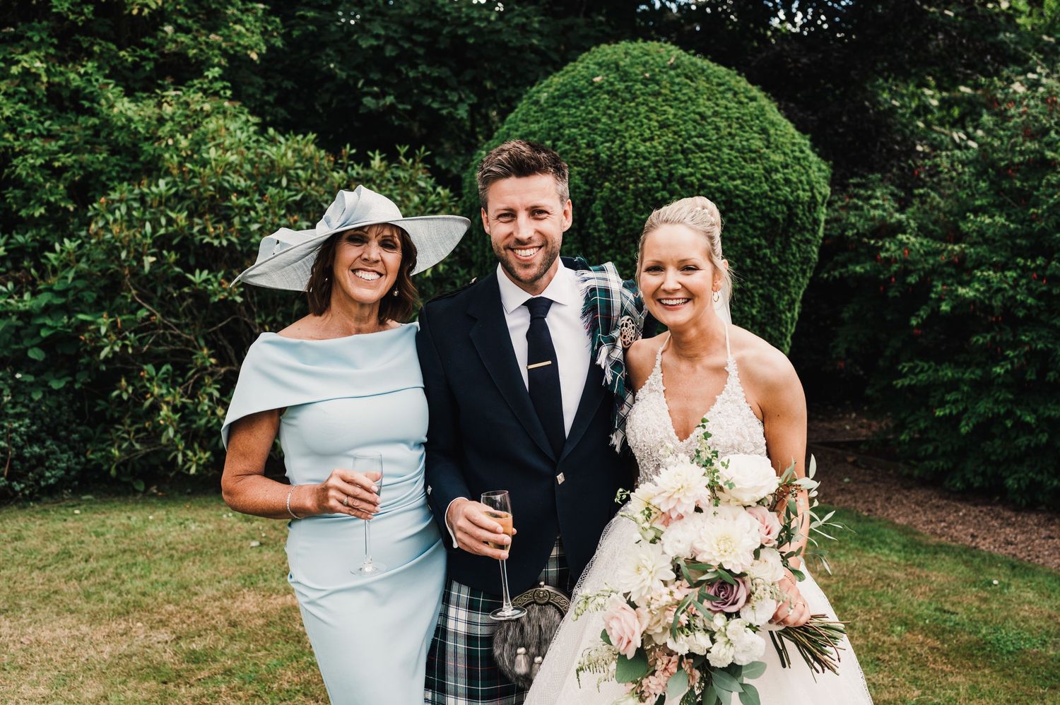 Wedding party members pose together on grass with round topiary bush in background.