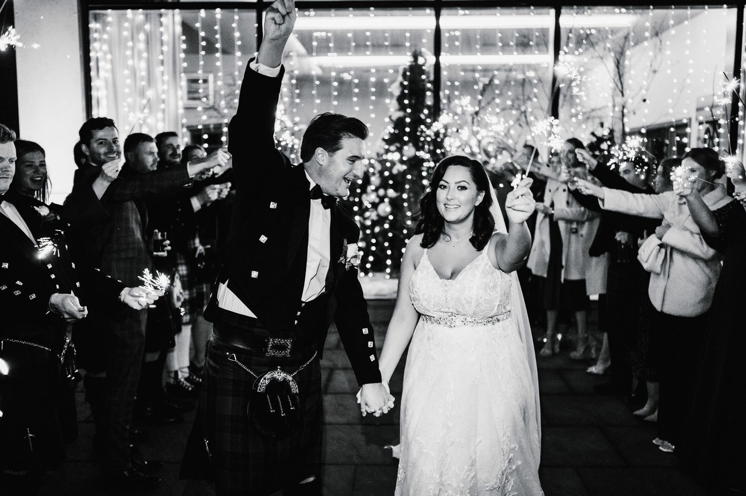 Joyful couple walks through sparkler exit tunnel at wedding reception with guests cheering.