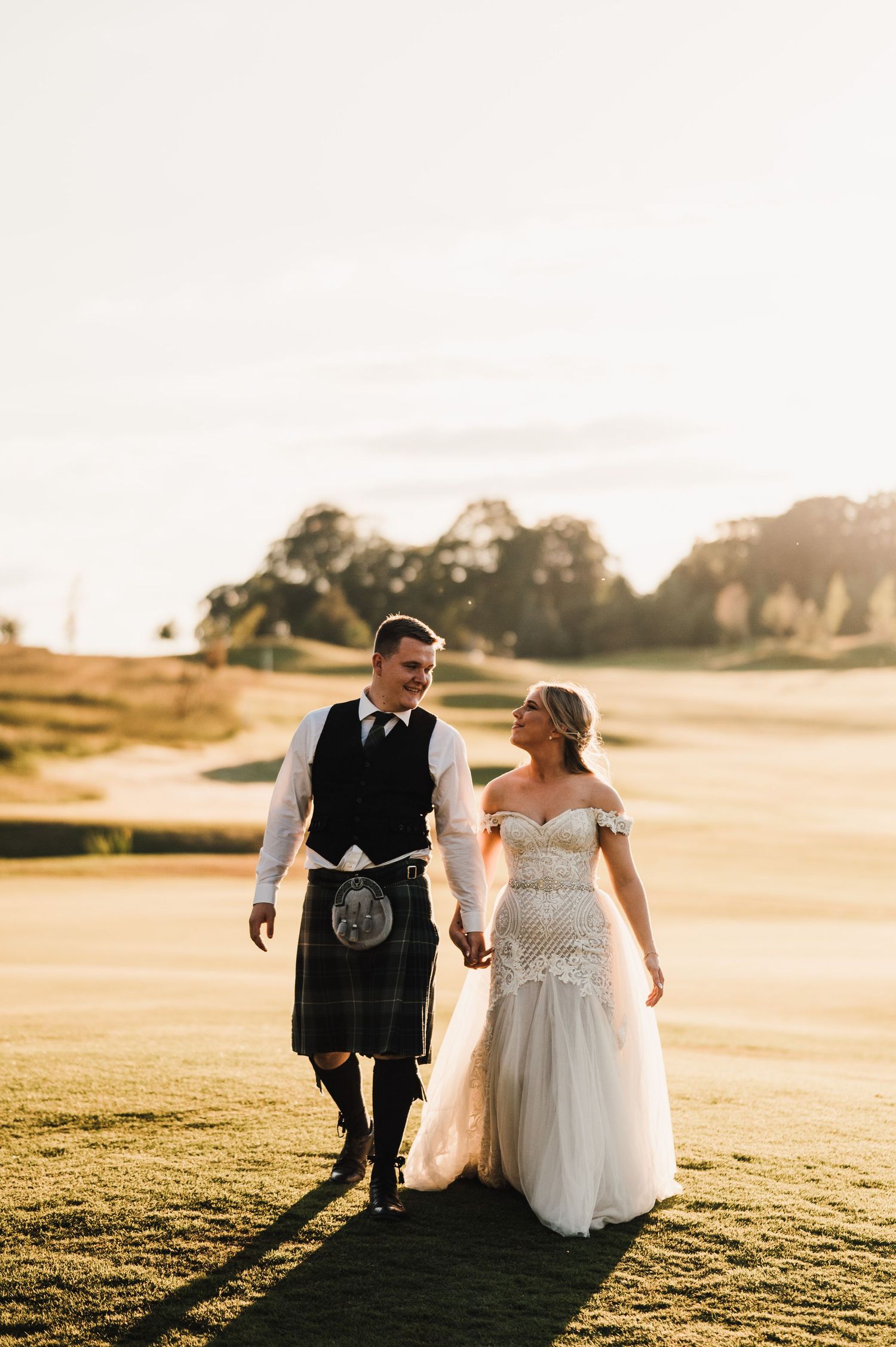 A couple in wedding attire walks hand in hand across a sun-lit golf course.