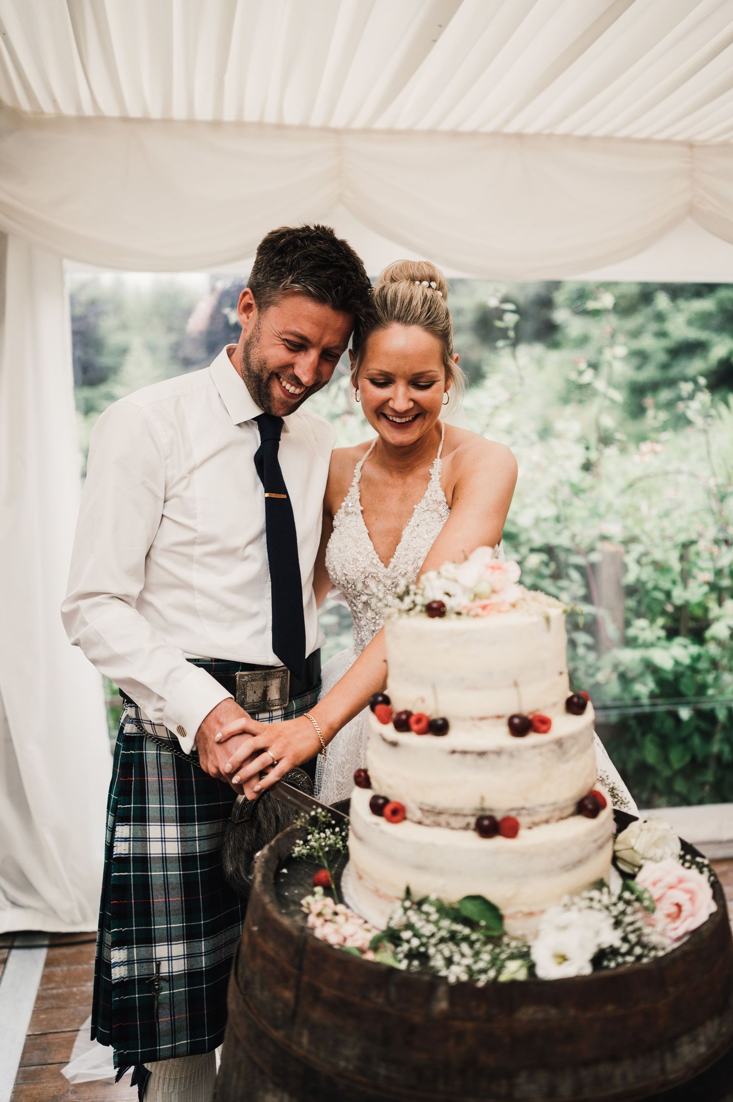 Wedding couple cutting their elegant three-tiered white cake decorated with berries in a draped tent setting.