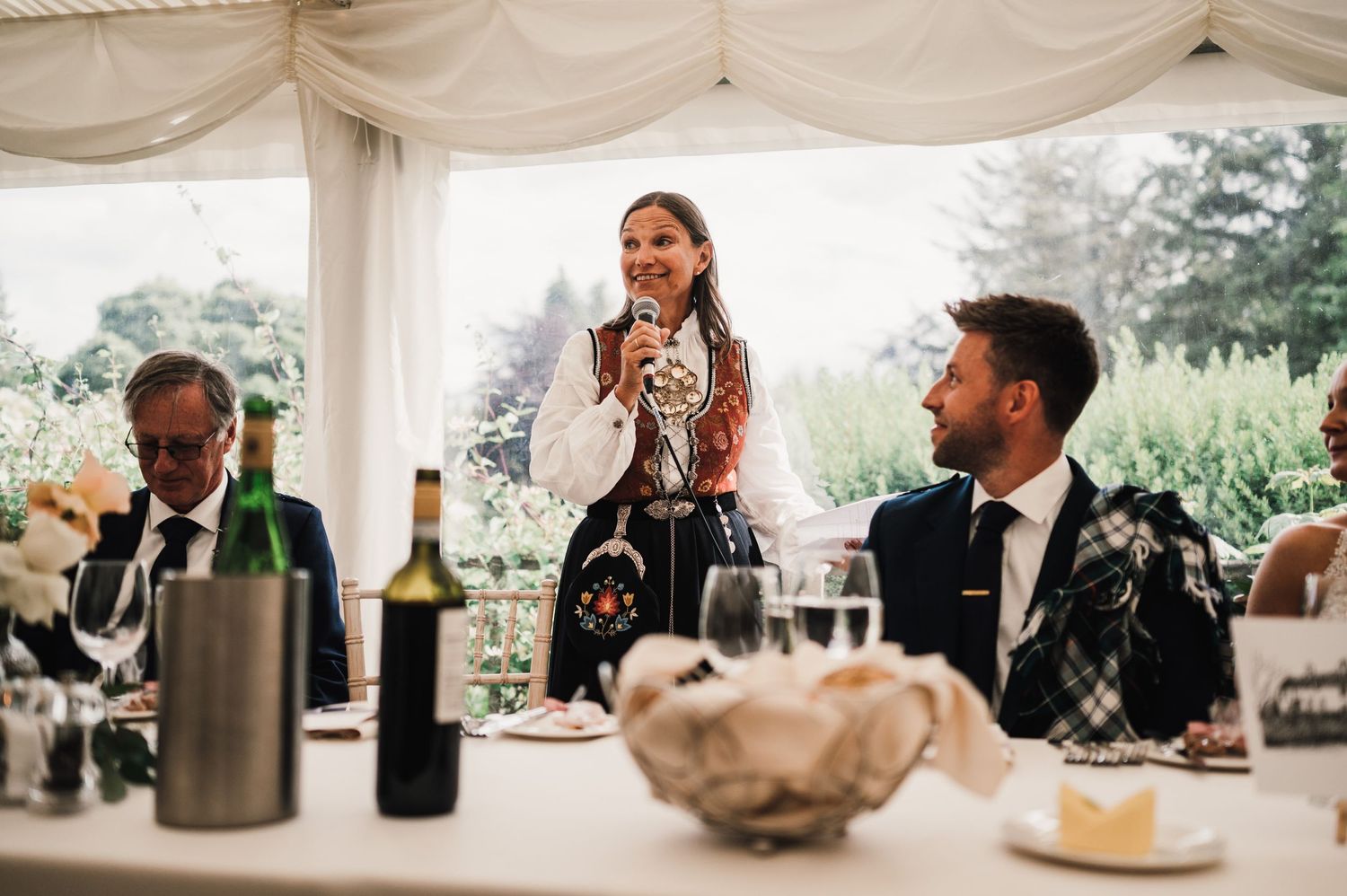 Wedding guests enjoy speeches and toasts under an elegant white tent with wine bottles on the table.