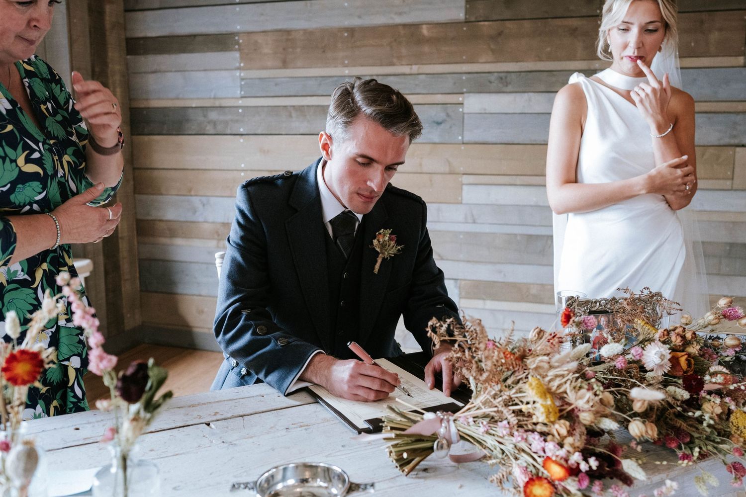 Newlyweds signing marriage documents together at rustic wooden table.