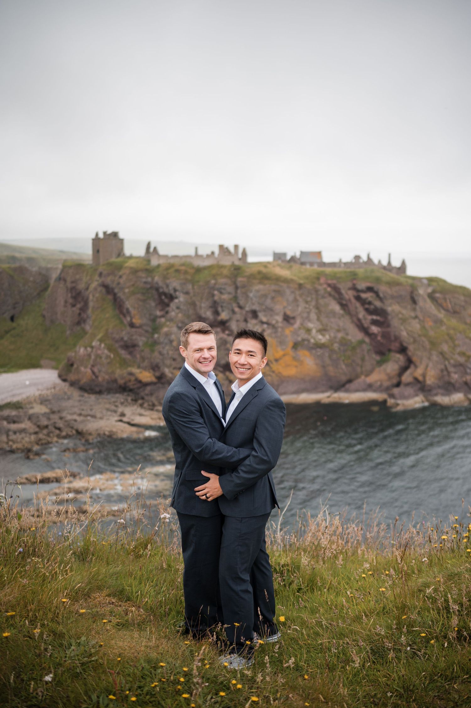 Same Sex Wedding Photography. Two people embrace on a scenic coastal hillside with ancient castle ruins visible in the misty background.