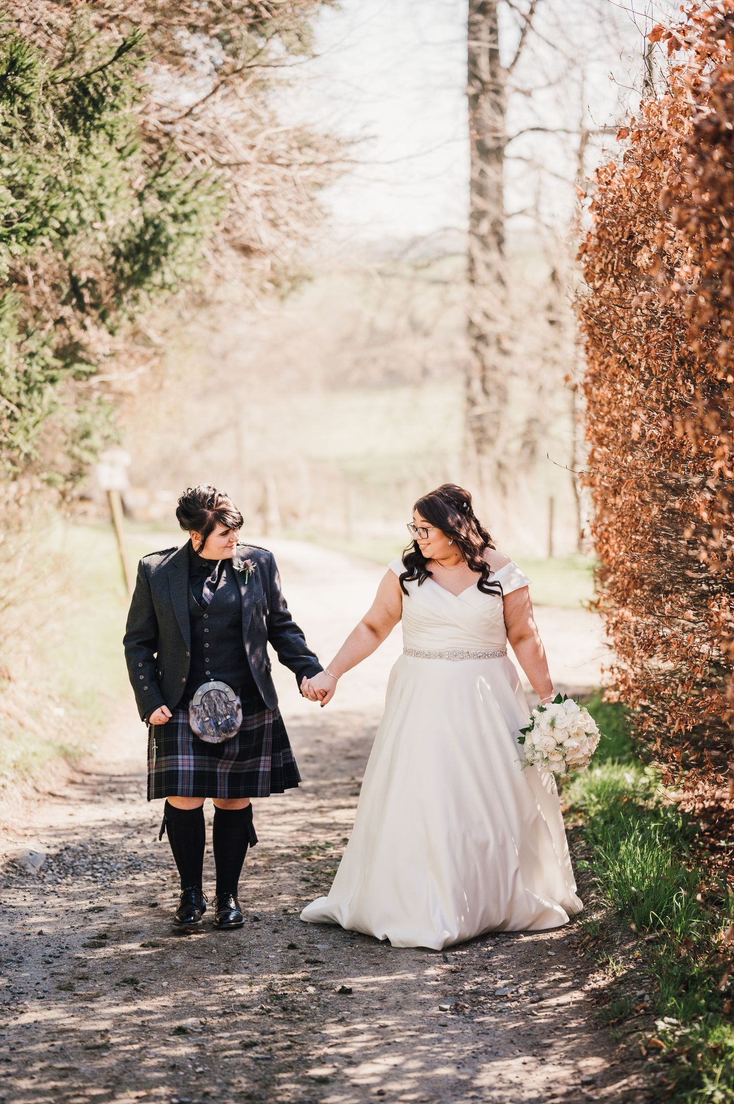 Logie HouA couple walks hand in hand on a woodland path, with one wearing a traditional Scottish kilt and the other in a flowing white gown.