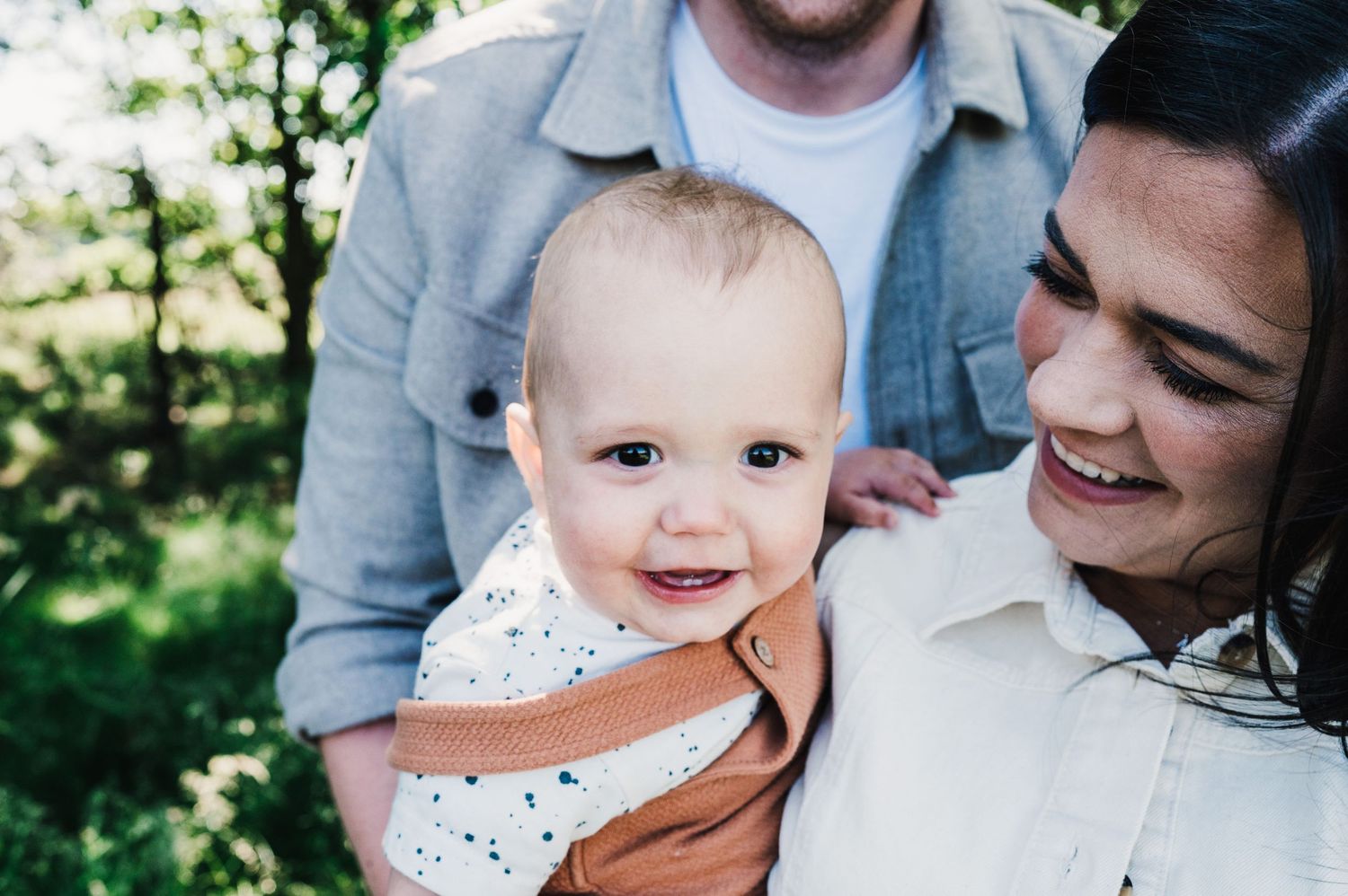 A baby wearing a brown leather backpack smiles while being held outdoors on a sunny day.