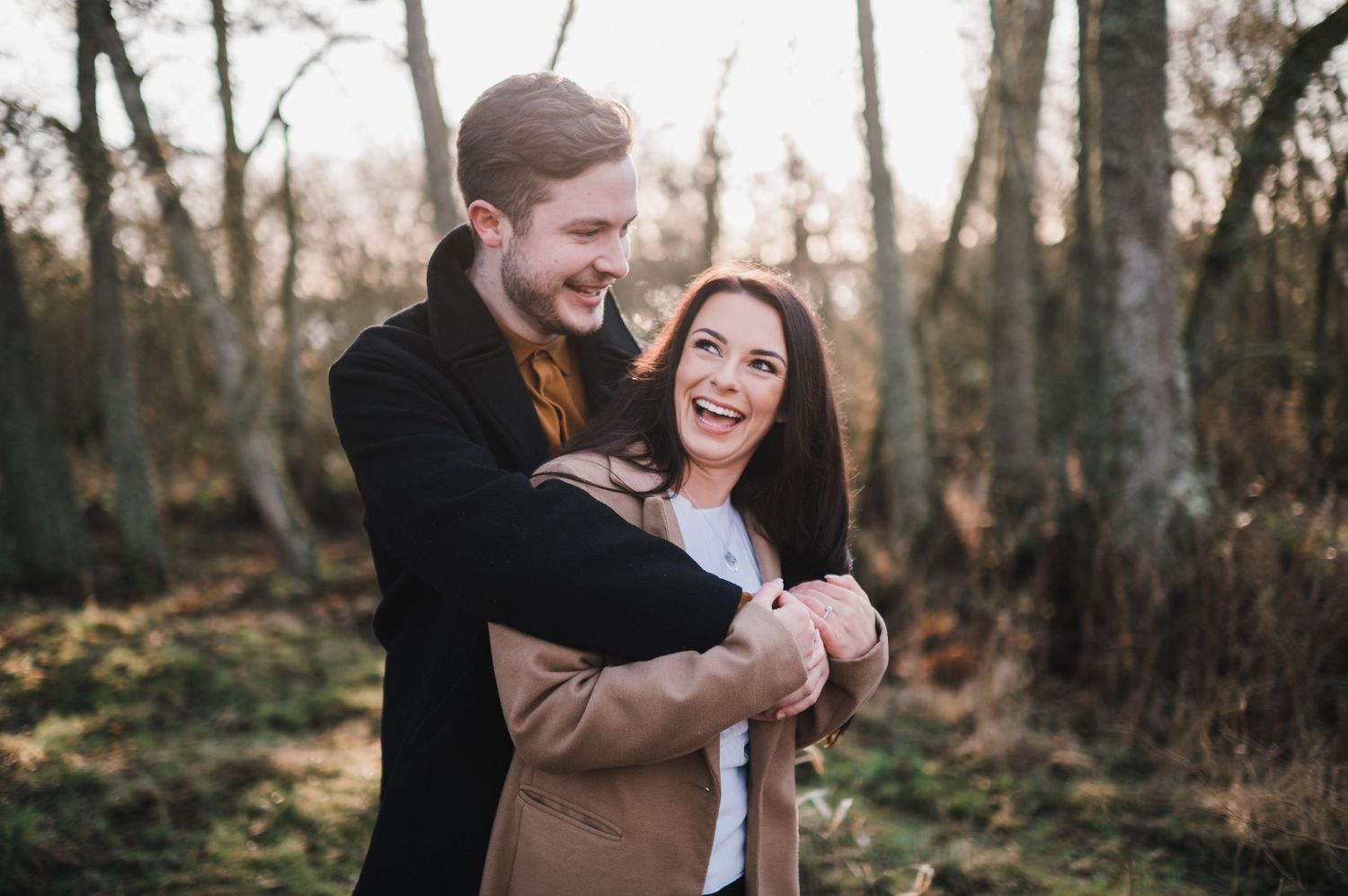 A couple embraces and laughs together in a wooded setting during golden hour.