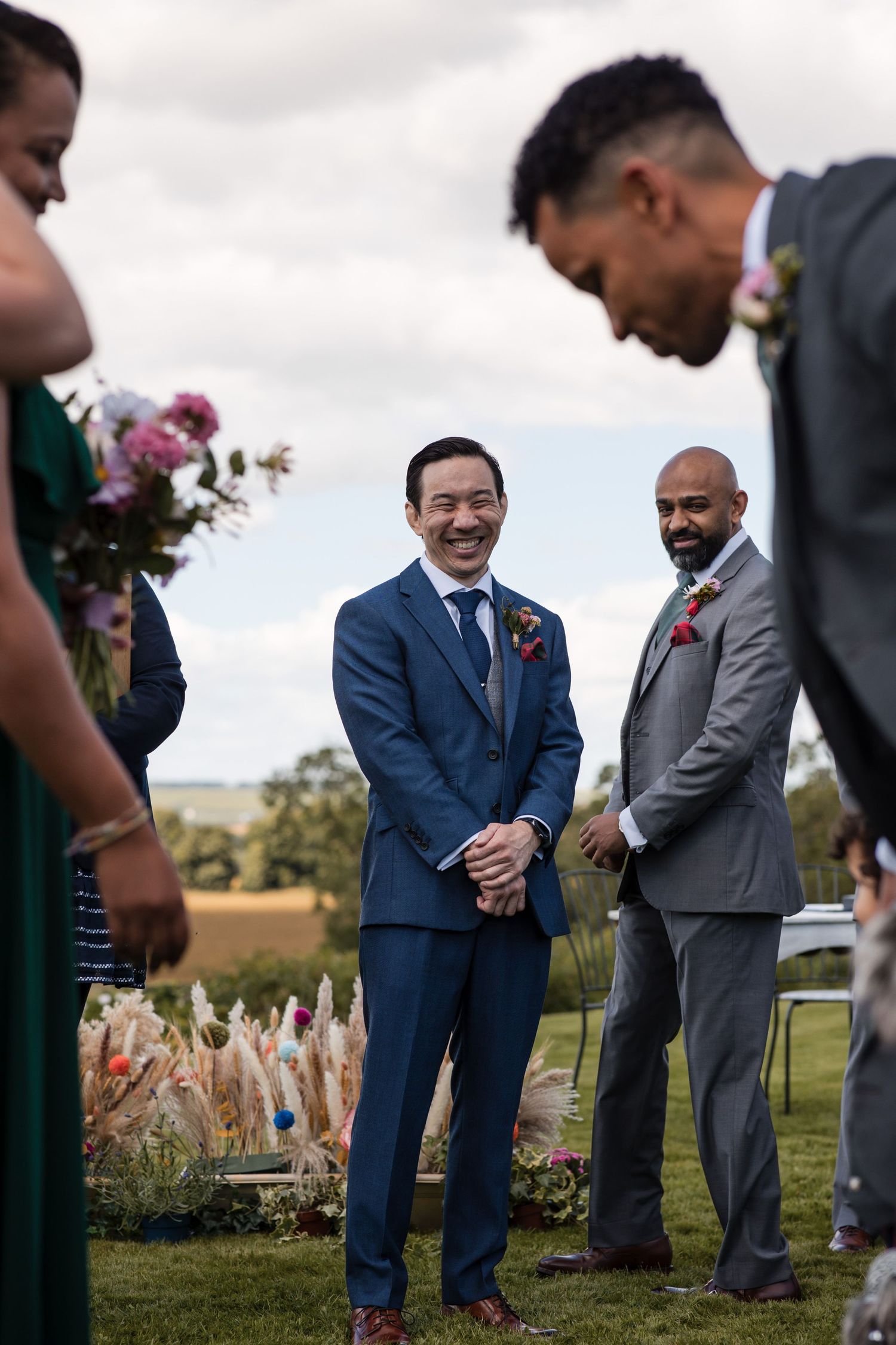 Wedding party members share a candid moment of laughter on a grassy lawn during an outdoor ceremony.