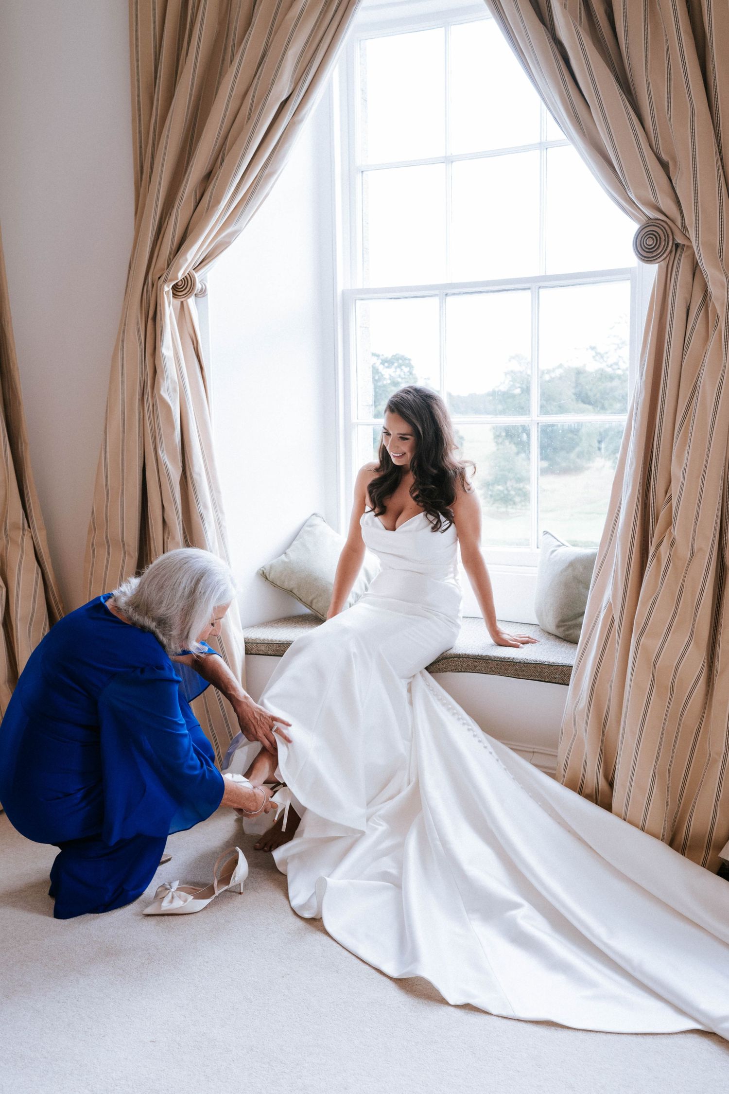 A wedding dress being adjusted near a window with elegant beige curtains.