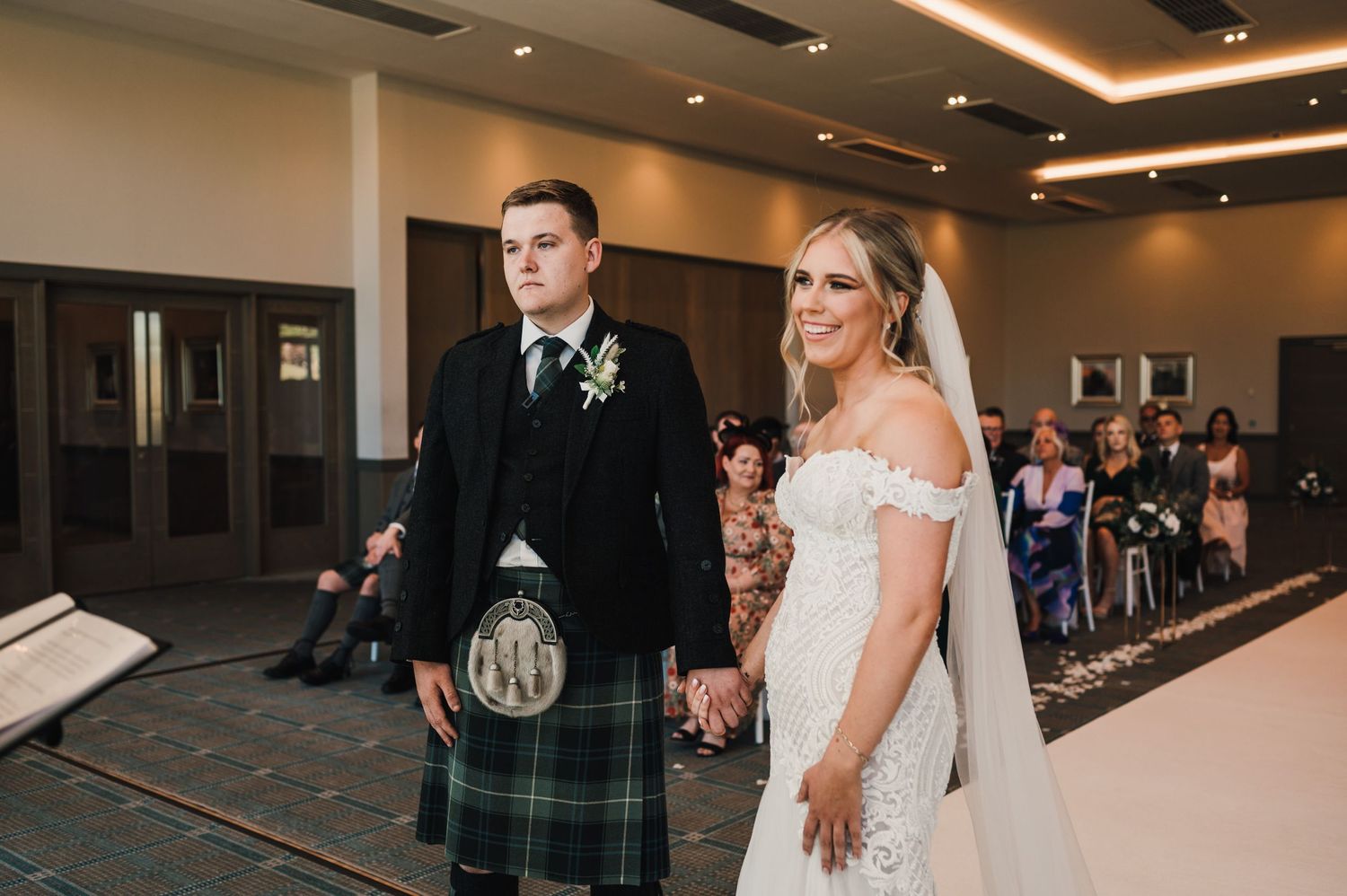 A Scottish wedding ceremony with a groom in traditional tartan kilt and bride in white dress walking down the aisle.