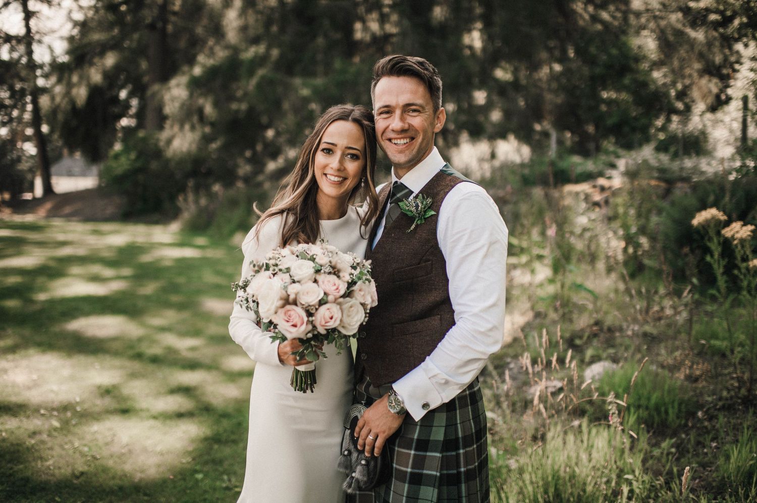 Wedding portrait of couple in forest with Scottish kilt and white dress holding pink floral bouquet.