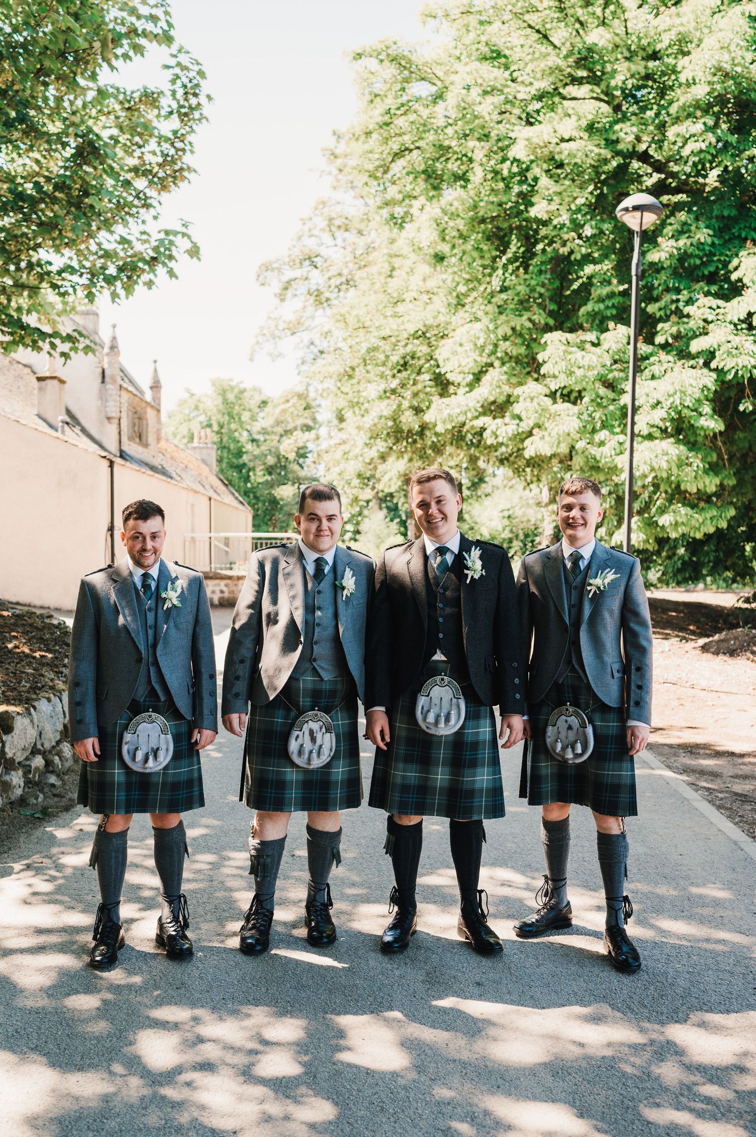 A wedding party in matching kilts and jackets poses on a sunny pathway.