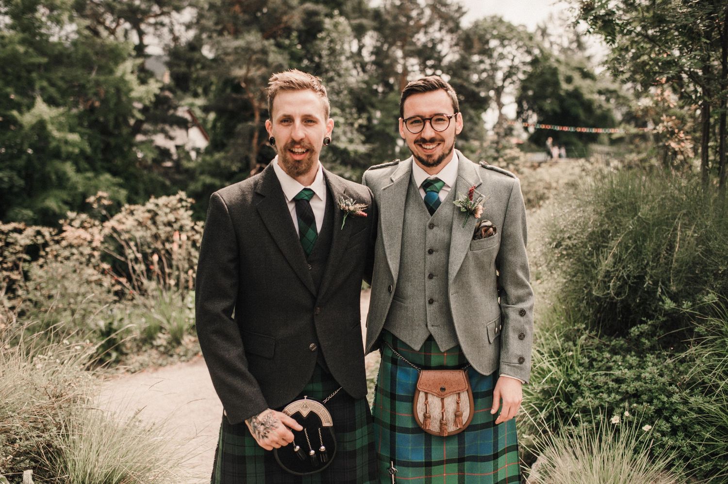 Two people in formal attire stand together outdoors, one wearing a traditional Scottish kilt and gray suit jacket.