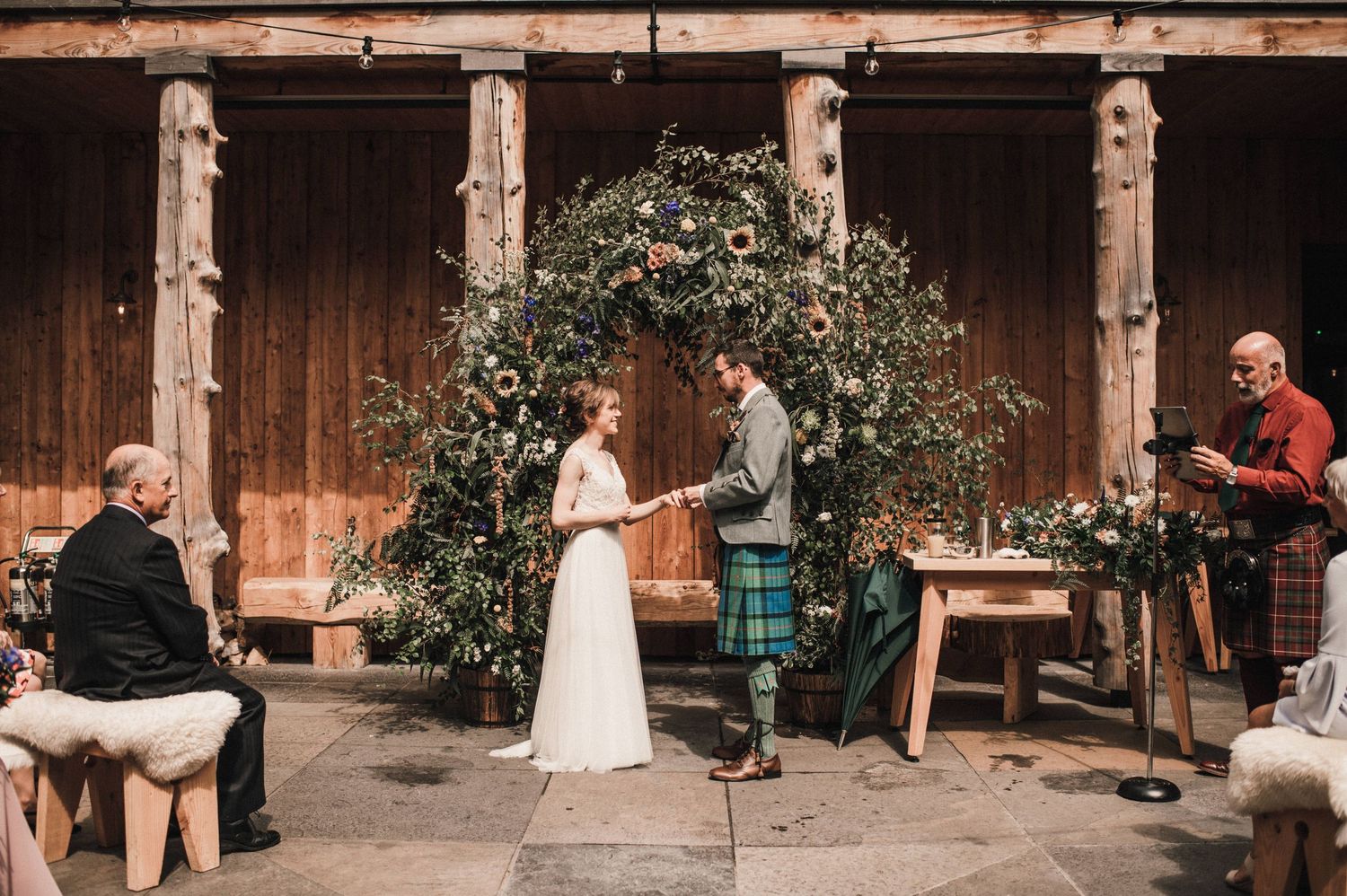 A small wedding ceremony taking place under a rustic wooden pergola with floral decorations.