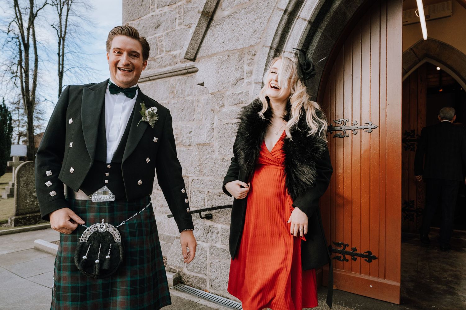 Couple in formal Scottish wedding attire smiling outside stone church door.