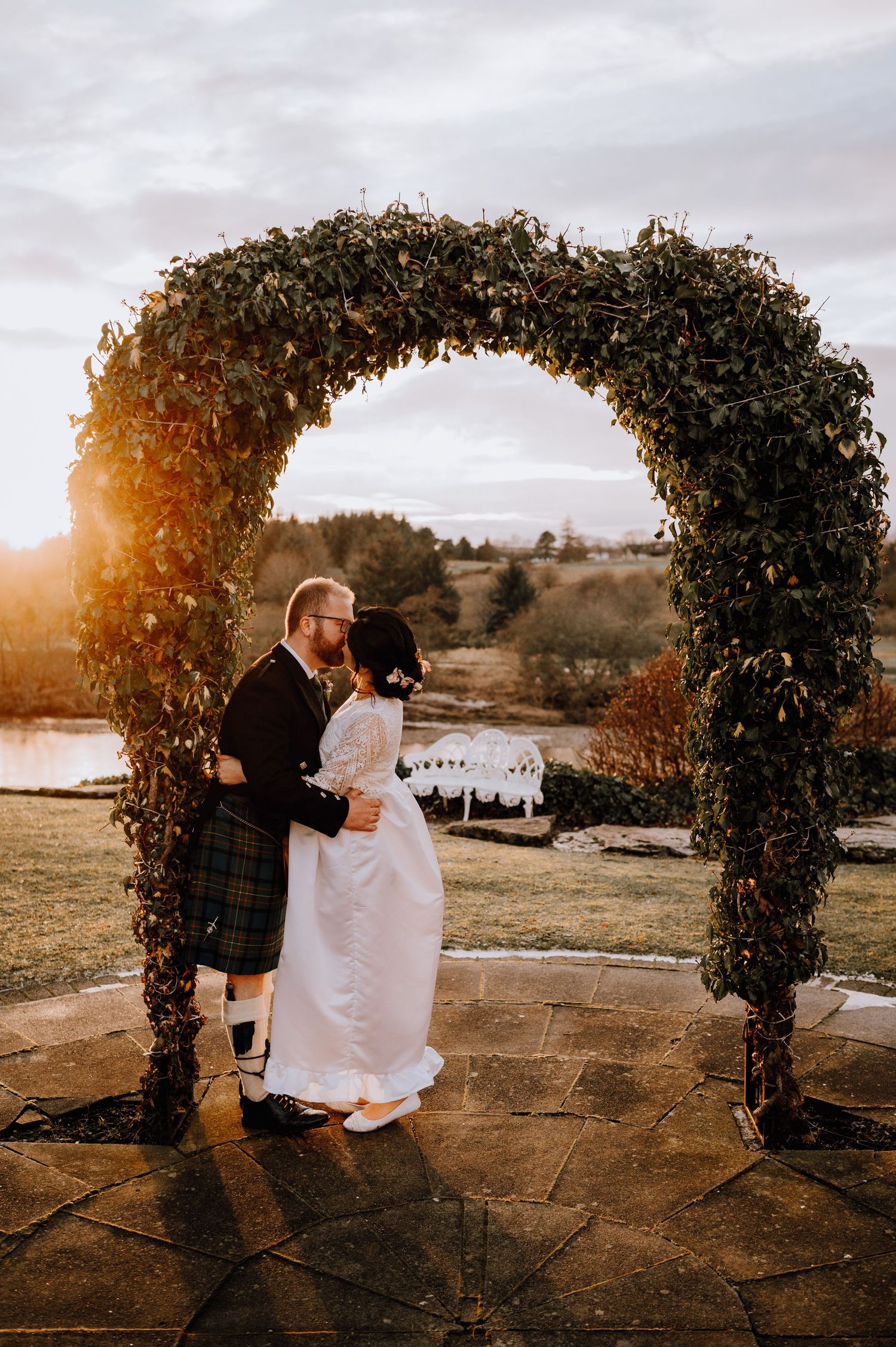 A couple shares an intimate embrace beneath a rustic garden arch during sunset at their Scottish-themed wedding.