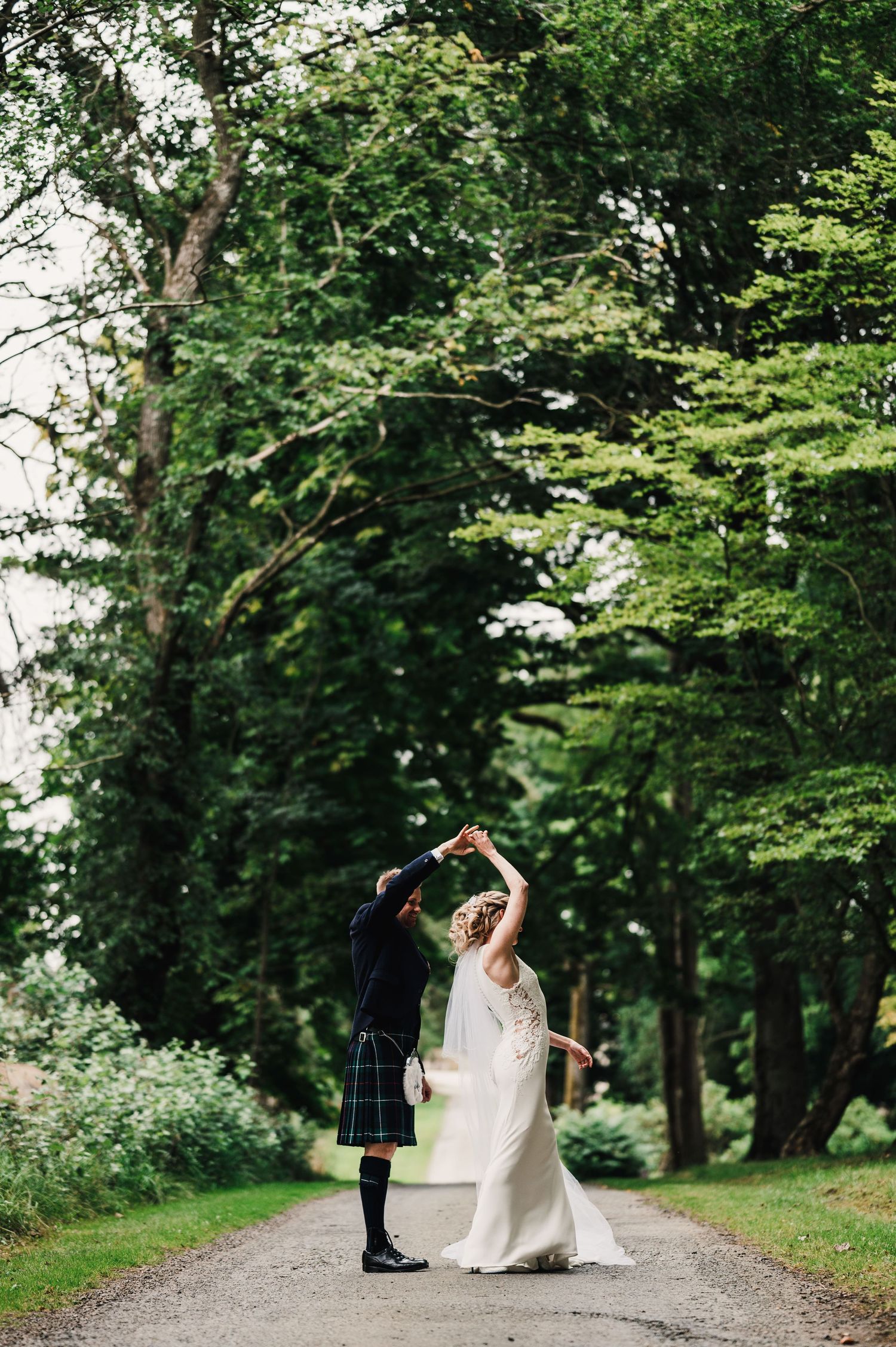 Couple dancing on a forest path, with the groom in traditional Scottish kilt and bride in elegant white dress.