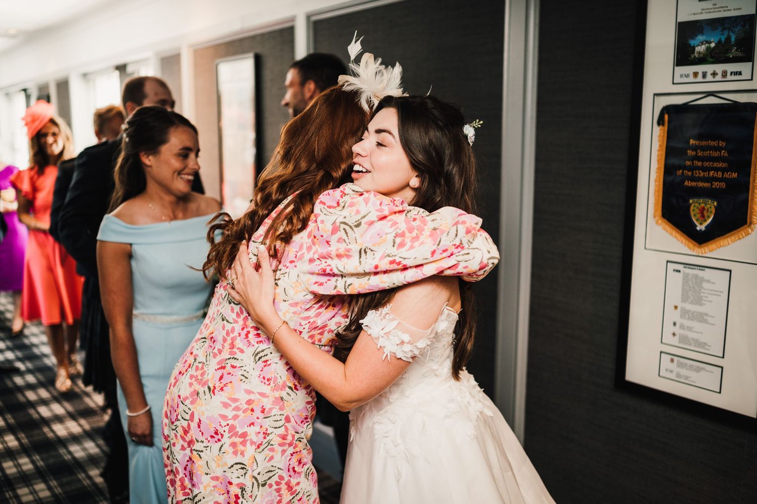 Two guests share a warm embrace at a wedding celebration in a hallway.