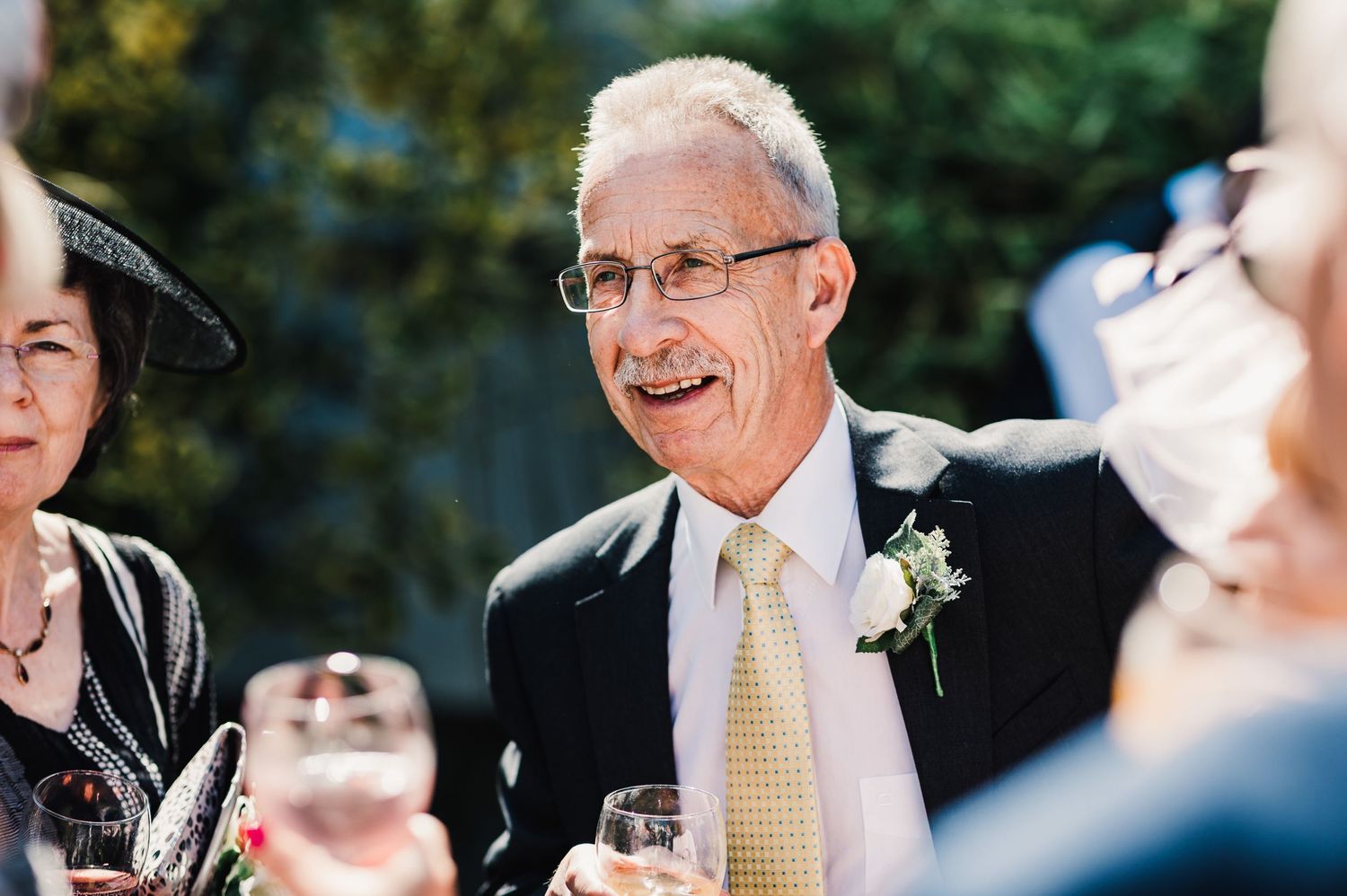 Wedding guest in formal attire smiling while holding champagne glass outdoors.