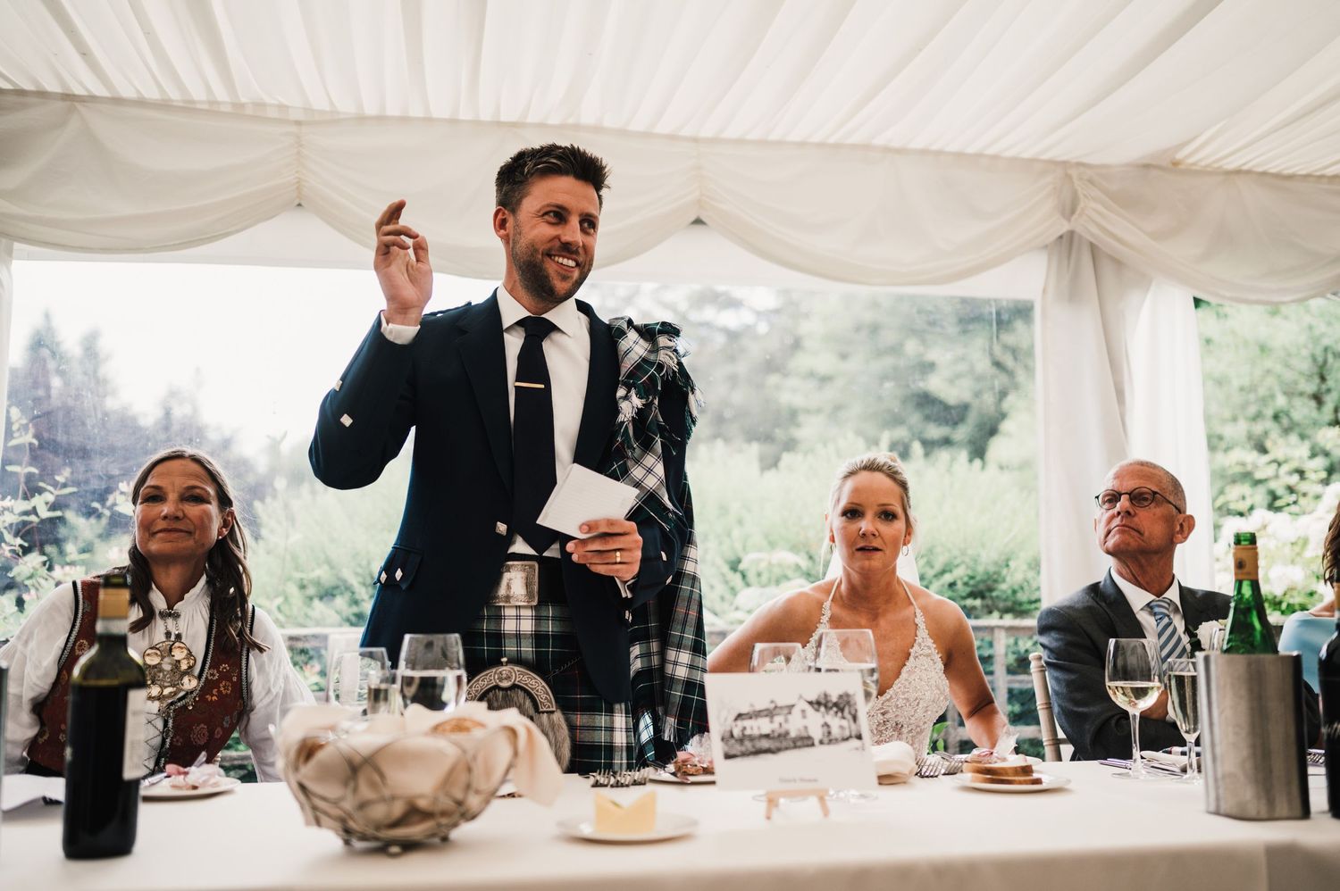 Speech being given at a Scottish wedding reception with traditional kilts and formal attire.