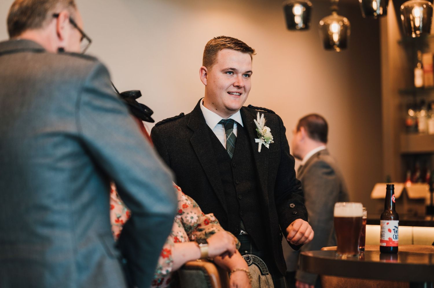 Wedding guest in black suit and boutonniere engaging in conversation at reception.