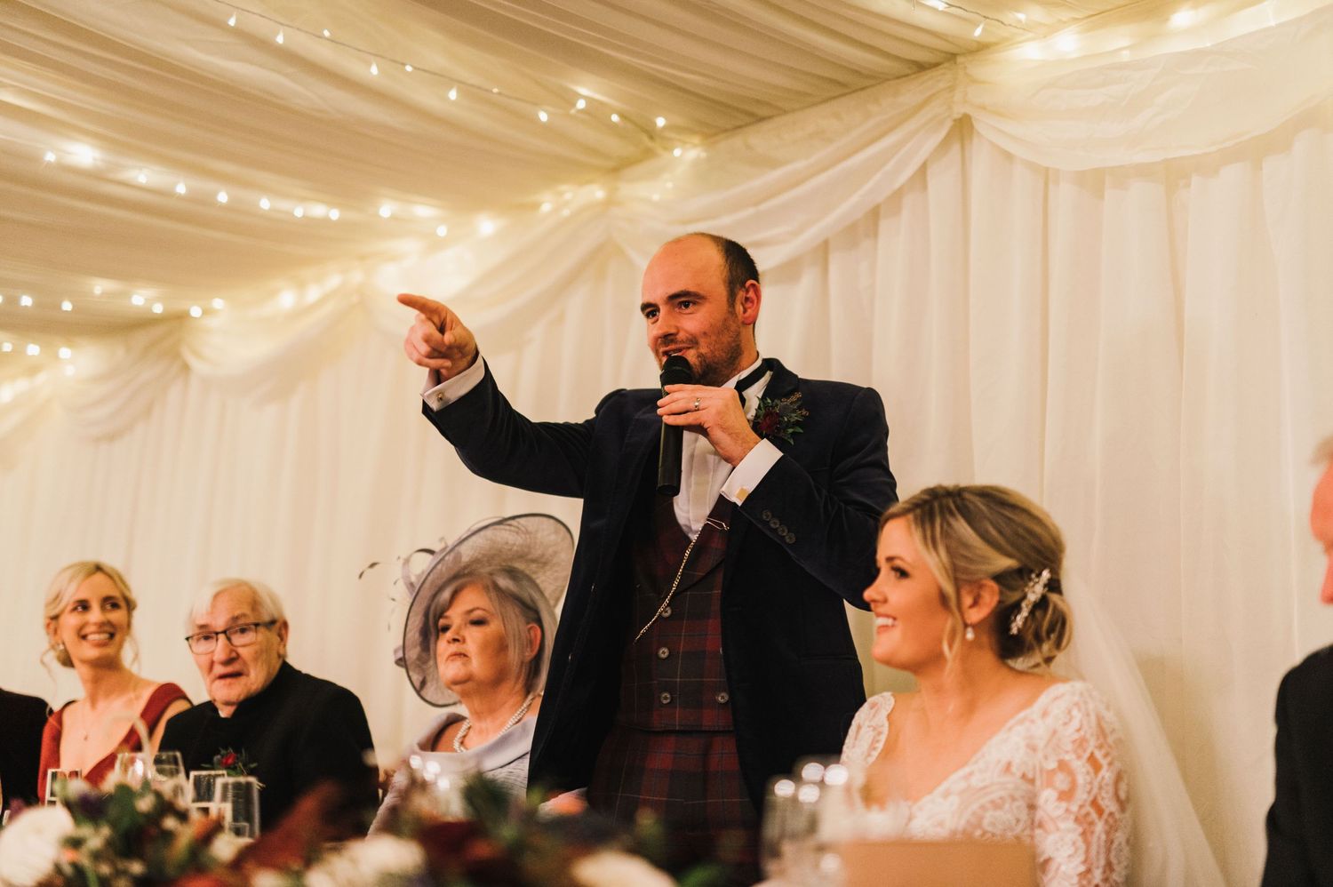 A man in formal wear gives an animated wedding speech while standing at the head table.