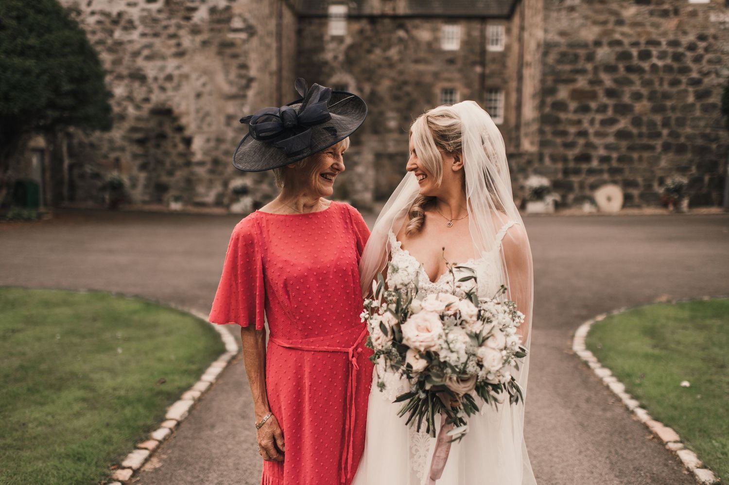 Candid moment between wedding guests in front of historic stone building with beautiful bouquet.